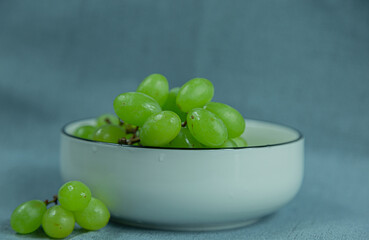 Against a solid color background, the white ceramic bowl is filled with fresh, tasty green grapes, with a scattering of a few grapes sitting next to the bowl.