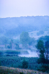 A cold morning landscape of a foggy lake surrounded by fields and hills with trees and tall grass.