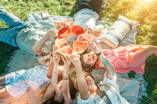 ..Young daughters with parents family lying on picnic blanket during weekend sunny day, smiling, laughing and rose up red juicy watermelon pieces. Family values, fruits vitamins, outdoor time concept