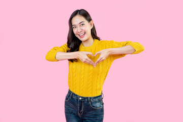 Young Asian woman feels happy and romantic shapes heart gesture expresses tender feelings wears casual jumper poses against pink background. People affection and care concept
