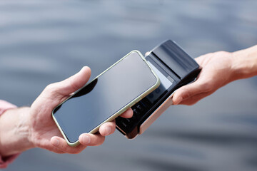 Closeup of unrecognizable person paying via NFC by smartphone outdoors with water in background,...