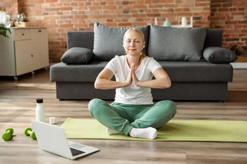 Gordijnen Mature woman sitting in yoga lotus position and doing breathing relaxation exercises, watching online lessons on laptop, workout in living room at home. Sport, fitness and healthy lifestyle concept © Oksana Klymenko