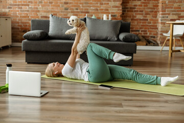 Mature woman doing sports exercises on mat with her pet, watching tutorial training videos of...
