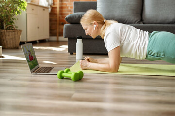 Senior woman on yoga mat doing yoga plank and watching online classes on laptop, workout in living...
