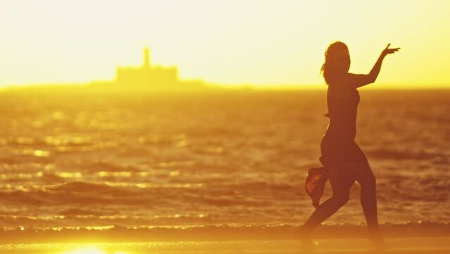 Graceful young woman in a dress dancing alone on the shore at bright orange sunset