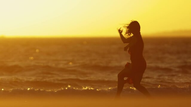 Graceful young woman in a dress dancing on the seashore at bright orange sunset