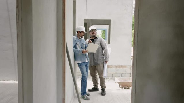 Indian architect with a mustache leans against the cement wall of an unfinished house. Talk to foreman engineer. Check plans for subordinates and design laptop computer houses under construction.