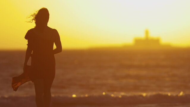 A young woman walking on the shore with bare feet at orange sunset