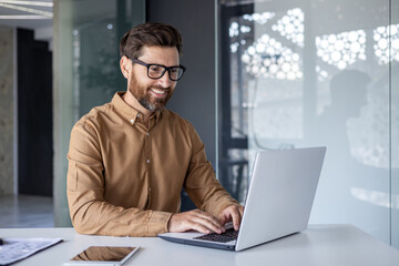 Experienced programmer inside office at workplace, senior man with beard and glasses working on laptop, businessman satisfied with achievement results