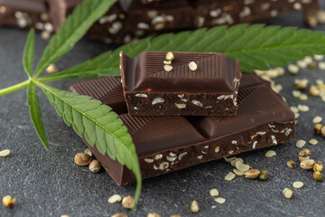 Broken dark chocolate bar with infused crushed hemp seeds on stone background. Vegan chocolate pieces and cannabis leaf and seeds closeup on dark table.