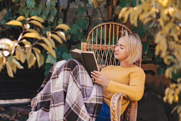 Beautiful girl resting sitting in autumn garden in chair wrapped in a woolen plaid blanket. Girl...