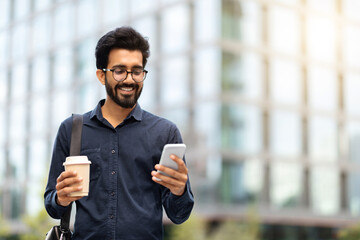 Cheerful indian guy going to office, using phone, drinking coffee