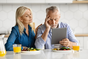 Upset senior spouses using digital tablet while eating breakfast in kitchen