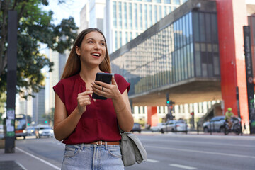 Happy business woman looking away with her smartphone walking on Paulista Avenue, Sao Paulo, Brazil