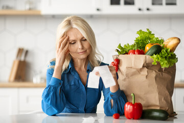 Grocery Expenses. Stressed Senior Lady In Kitchen Checking Bill After Food Shopping