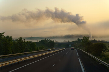 Near the motorway there is an industrial enterprise from the chimney of which there is thick smoke, in the rays of the rising sun