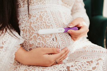 Pregnant woman sitting strokes hugging the belly tummy abdomen, hold pregnancy test kit. Future family, baby infant expecting child inside, lady in white dress
