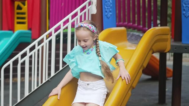 Smiling little girl rides plastic slide in maze on safe children playground happy child with braids plays attraction in public urban park schoolgirl enjoys sliding