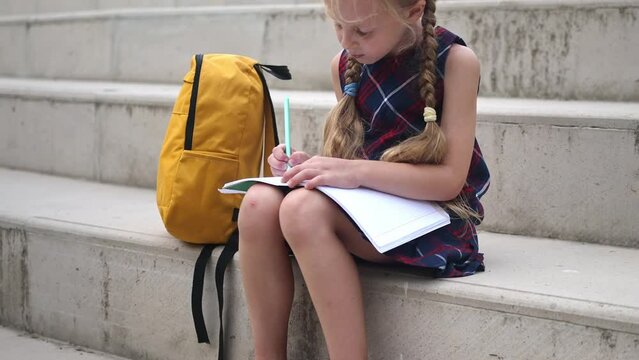 Adorable little girl draws in exercise book sitting by rucksack on stone stairs blonde schoolgirl with plaits does homework in school yard primary school student