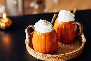 Pumpkin shaped cups of hot drink withwith whipped cream, cinnamon stick on black wooden table in cozy interior. Pumpkin latte. Cozy autumn beverage for Thanksgiving, Halloween holidays at home.