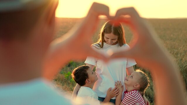 Mom Hugs Little Children, Dad Looks At Them Through Heart Sign In Park. Love For Family For Child And Mother. Looking Through Heart Symbol Made By Fingers At Happy Family. Mom Dad Child. Son Daughter