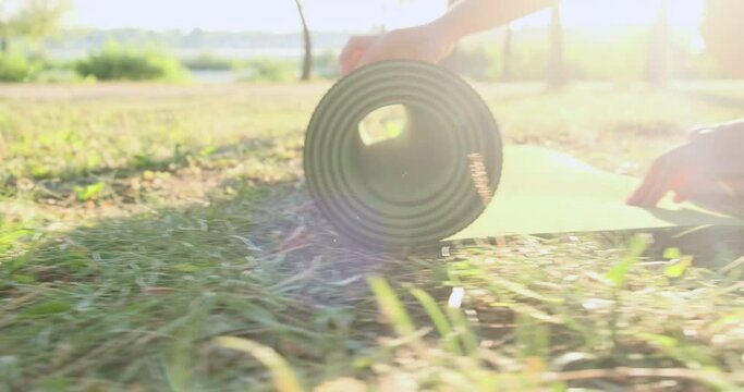 Yoga mat slowly unfolds on green lawn in city park, side view, close-up, panning, slow motion. Woman's hands let go of yoga mat