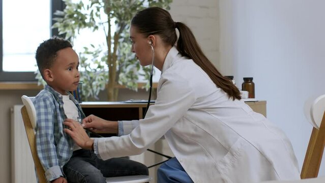 Woman pediatrician listening to little african american patient lungs at clinic and writing result in medical history