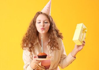 Beautiful woman with Birthday gift blowing out candle on cake against yellow background