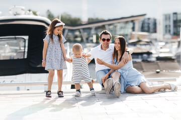 A family of four walks in the marina with yachts in the background. Mom and dad together with their little son and daughter. Happy family spending leisure time outdoors