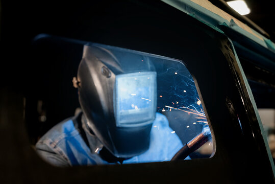 A professional welder works using welding equipment and makes seams on metal. The welder is wearing a protective mask and gloves. Sparks and fire when using welding equipment