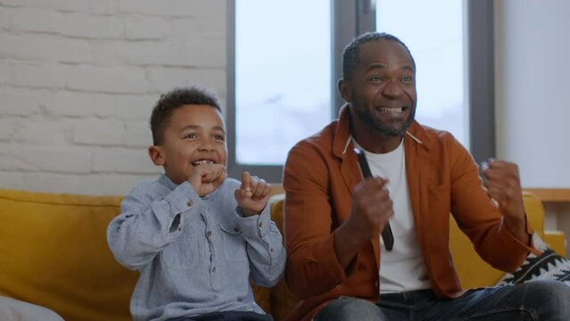 Excited emotional african american dad and son watching football game on tv, enjoying goal, tracking shot, slow motion