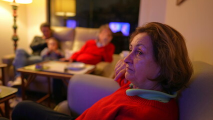 Senior woman watching television seated on couch, family in background. Boomer generation lady...