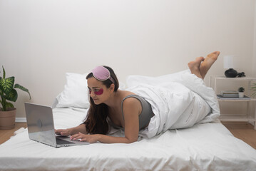 Young lady lies on bed in bedroom looking at laptop screen with smile woman with sleeping mask and patches rests before going to bed browsing Internet