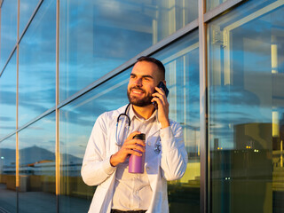 young male doctor talking by phone outdoors in front of a hospital with a coffee thermos on his hand
