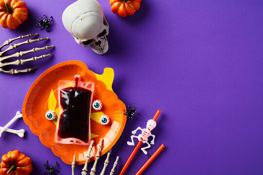 Happy Halloween holiday concept. Halloween table with pumpkin plate, blood packet, skeleton hands, skull, pumpkins on purple background. Top view, overhead.