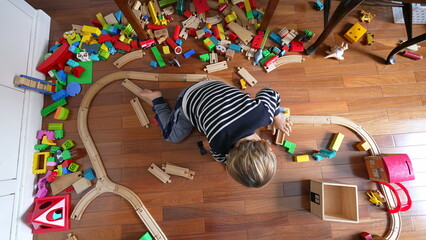 Small boy playing with toys seen from above perspective, child immersed in play with retro vintage railroad tracks and blocks scattered around - Powered by Adobe