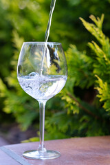 A clear glass wine glass stands on a table in the garden with a stream of water pouring into it.