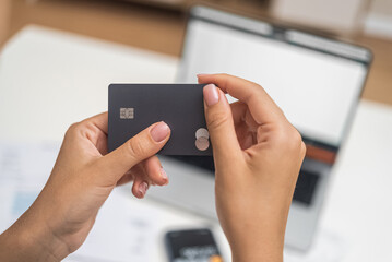 Female hands hold dark credit card for online payment at desk with documents at home woman plans payment in banking system on blurred laptop background