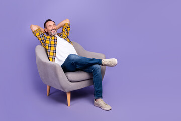 Photo of satisfied positive man hold arms behind head relax comfort trendy furniture empty space isolated on purple color background