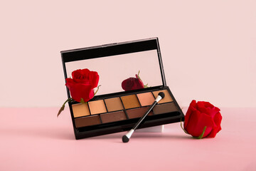 Palette of stylish eyeshadows, makeup brush and beautiful red rose flowers on pink background