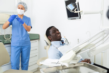 Positive african-american man patient sitting on dental chair and smiling while Asian woman dentist standing in background and preparing tools for examination.