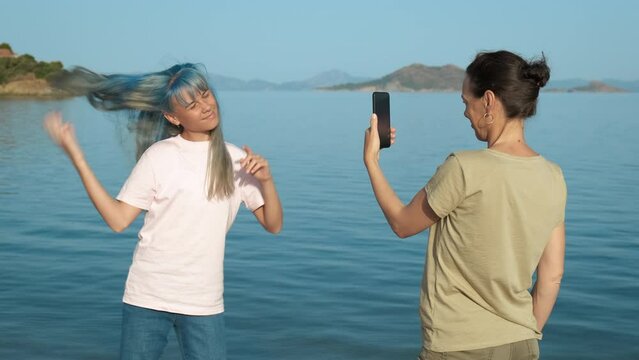 A young girl is dancing by the sea, her mother takes pictures on the phone.
