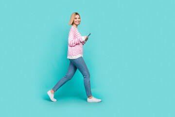 Full length photo of adorable cute girl dressed striped sweater walking texting gadget empty space isolated teal color background