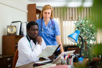 Portrait of two multinational doctors in the office of a resident of the clinic, sitting at a table...