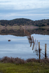 A wooden and wire fence is going across and large body of water. The water is blue. There are hills behind the water. The sky is light blue and cloudy. Vertical photo
