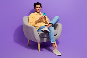 Fototapeta na wymiar Full body size photo of cheerful student guy sitting soft armchair browsing phone social medias retweet isolated on purple color background