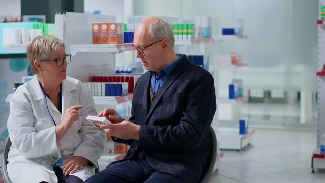 Proficient pharmacist in drugstore with patient, suggesting him heart arrhythmia medication pills to treat disease symptoms. Octogenarian man receiving treatment from employee