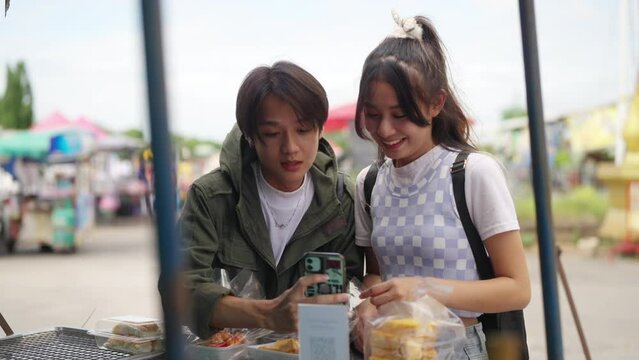 Group of Happy Asian people tourist buying and eating street food with using mobile app on smartphone scan QR code making online payment during travel local town in Thailand on summer holiday vacation