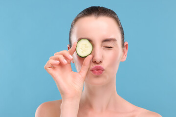 Beautiful woman covering eye with piece of cucumber and blowing kiss on light blue background