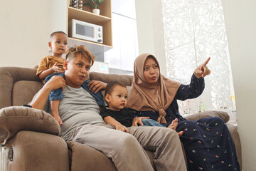 Happy Asian muslim family with two children sitting on couch at home watching tv together